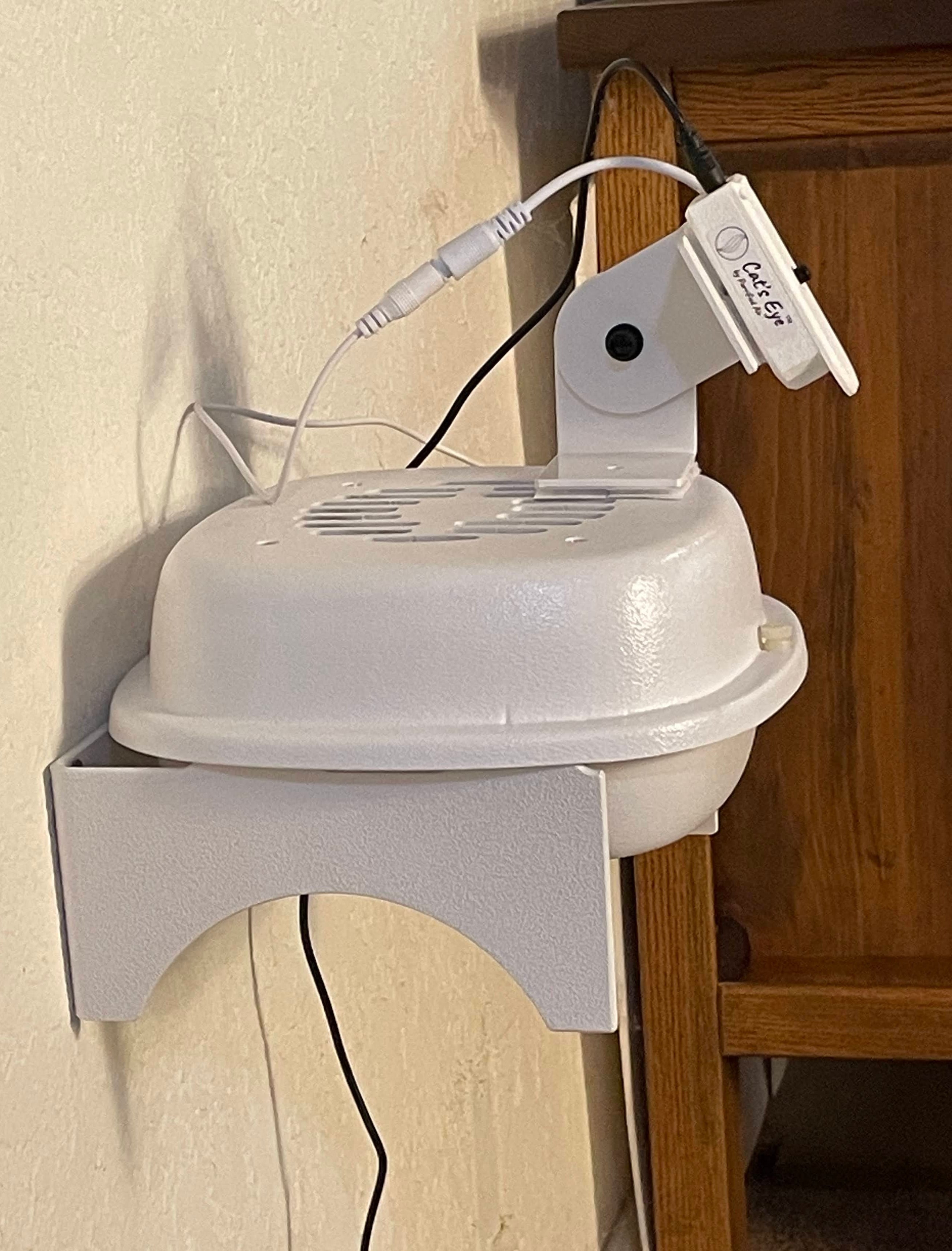A white circular fan or dryer-like appliance with a side support bracket and attached cord, photographed on a light floor in front of a wooden cabinet.