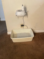 A white plastic cat litter box filled with gray litter, placed on a brown carpeted floor next to a beige wall with a mounted white device and power cord.