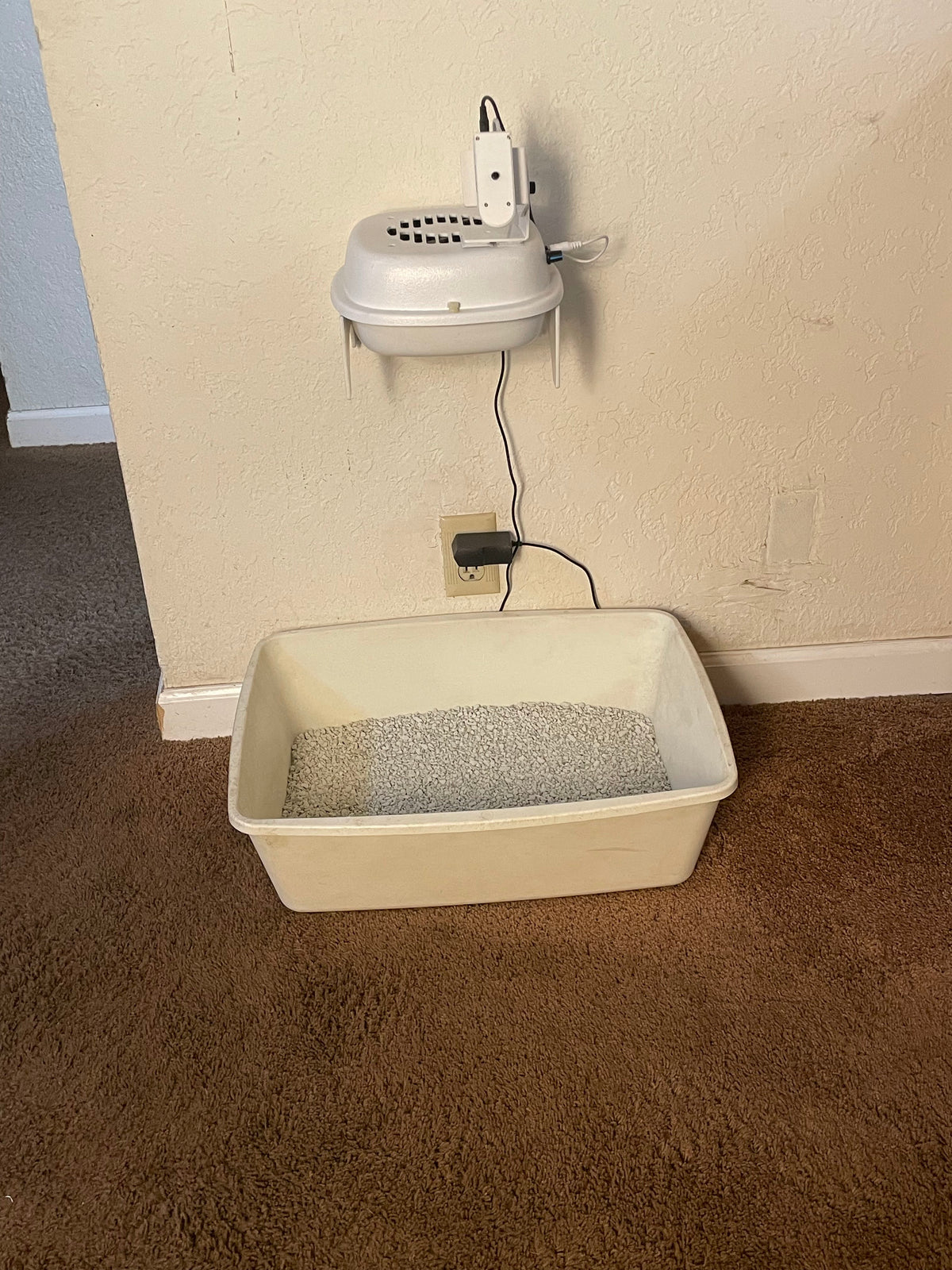 A white plastic cat litter box filled with gray litter, placed on a brown carpeted floor next to a beige wall with a mounted white device and power cord.